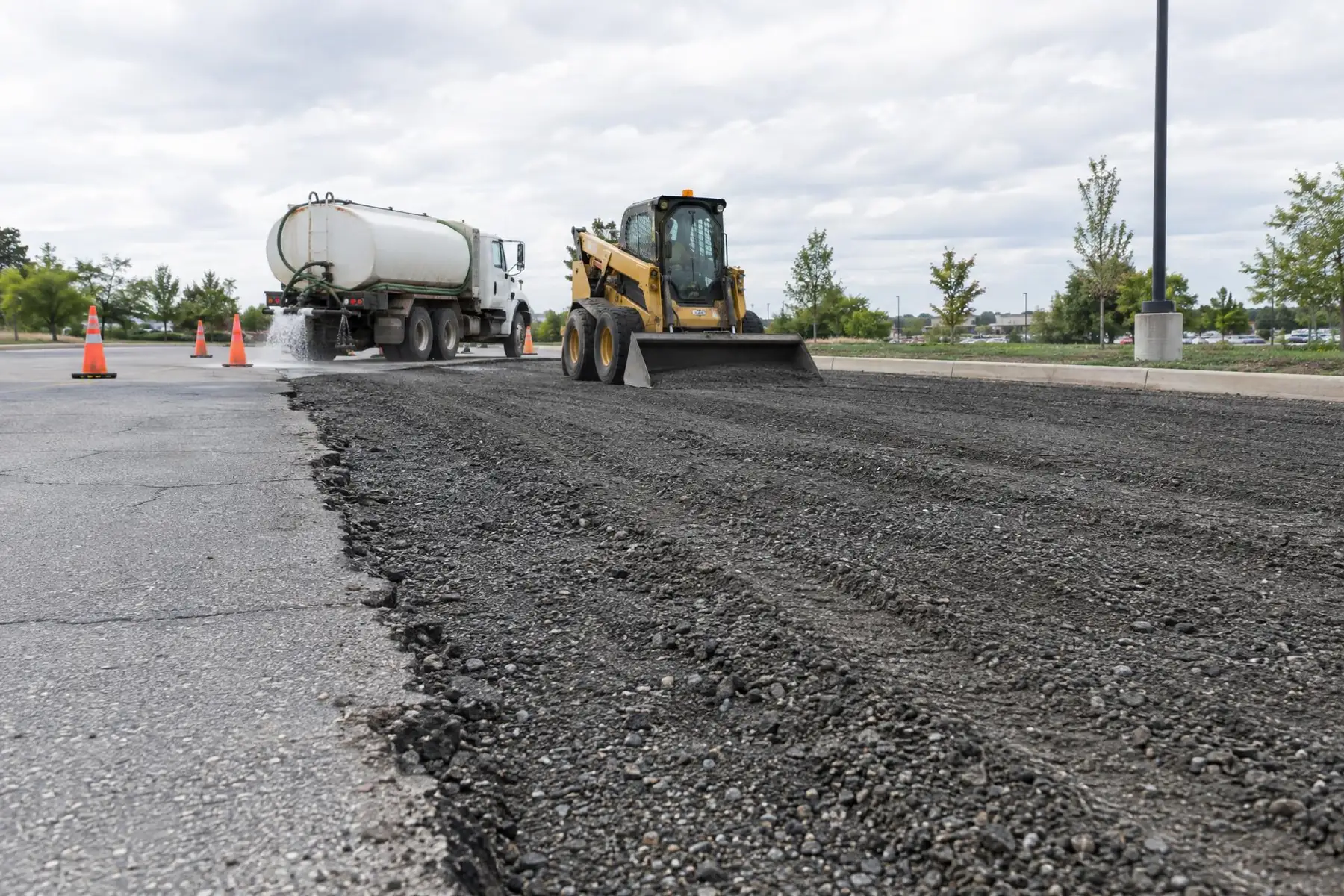 Parking lot with the old surface stripped to stone base while a skid steer and water truck work beside cones